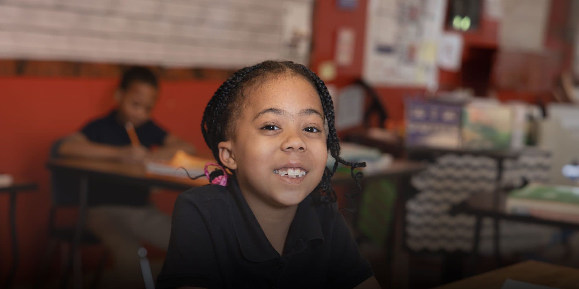 Smiling student with braided hair sitting at a desk in a classroom.