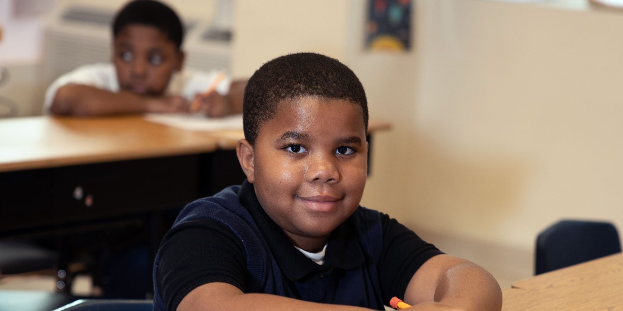 Smiling student in a navy polo sitting at a classroom desk.