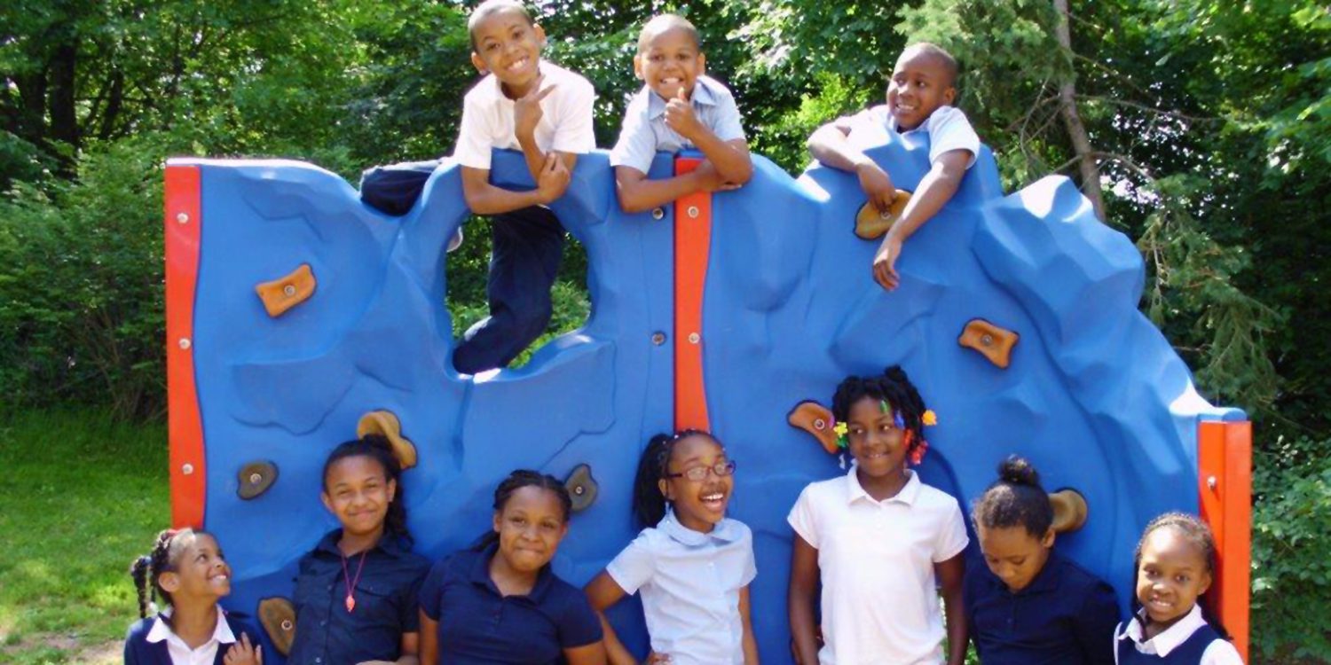 Elementary students standing outside by rock climbing playground structure.